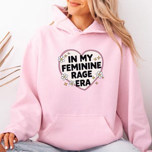 Könnte beinhalten: Ein hellrosa Kapuzenpullover mit einer zentralen Grafik in Form eines weißen Herzens mit rosa Umrandung. Im Inneren des Herzens steht der schwarze Text IN MY FEMININE RAGE ERA, umgeben von kleinen weißen Gänseblümchen und gelben Sternsymbolen.