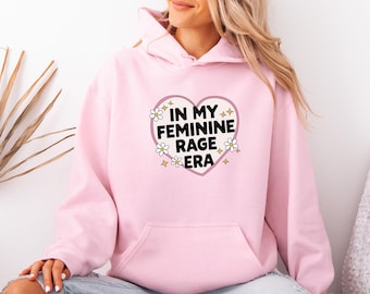 Könnte beinhalten: Ein hellrosa Kapuzenpullover mit einer zentralen Grafik in Form eines weißen Herzens mit rosa Umrandung. Im Inneren des Herzens steht der schwarze Text IN MY FEMININE RAGE ERA, umgeben von kleinen weißen Gänseblümchen und gelben Sternsymbolen.