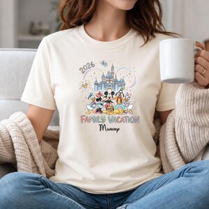 May include: Cream-colored t-shirt featuring a colorful graphic of Disney characters in front of a castle. The text "2026 Family Vacation Mummp" is printed below the image. The shirt is made of a soft material.