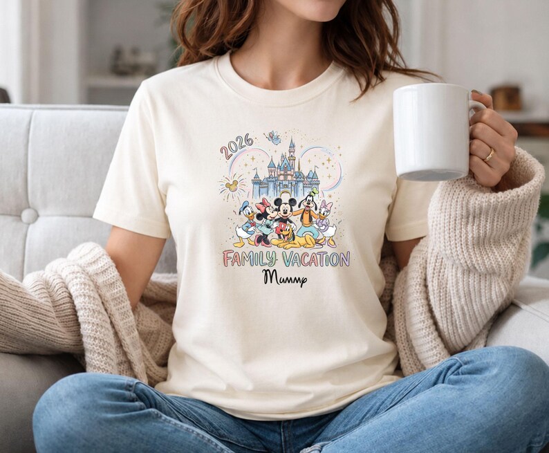 May include: Cream-colored t-shirt featuring a colorful graphic of Disney characters in front of a castle. The text "2026 Family Vacation Mummp" is printed below the image. The shirt is made of a soft material.