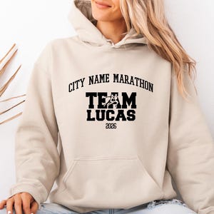 May include: A beige hooded sweatshirt featuring black text that reads CITY NAME MARATHON, TEAM LUCAS, and 2026. The design includes a graphic of two runners integrated into the word TEAM. The hoodie has long sleeves and a front kangaroo pocket.