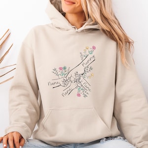 Op de afbeelding: Beige hoodie met een afbeelding van handen die elkaar vasthouden, met het woord "Mummy" en twee keer het woord "Name" in script. Het ontwerp bevat bloemaccenten in roze, geel en groen.