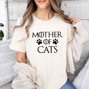 May include: A white t-shirt with the text "Mother of Cats" in black lettering. The text is styled to look like the "Game of Thrones" title card, with paw prints replacing the swords.
