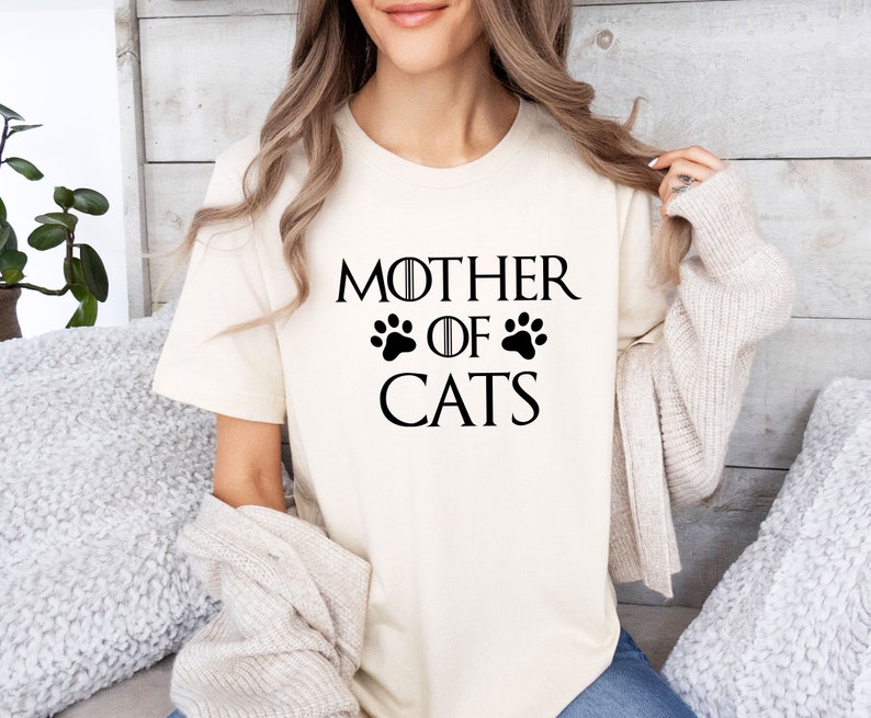 May include: A white t-shirt with the text "Mother of Cats" in black lettering. The text is styled to look like the "Game of Thrones" title card, with paw prints replacing the swords.
