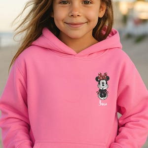 May include: A bright pink hooded sweatshirt featuring a Minnie Mouse graphic on the front. The graphic includes Minnie's face, bow, and outfit. The name "Isla" is printed below the graphic in white script.