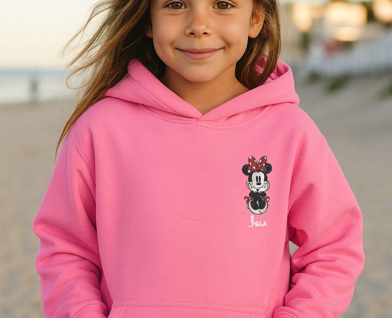 May include: A bright pink hooded sweatshirt featuring a Minnie Mouse graphic on the front. The graphic includes Minnie's face, bow, and outfit. The name "Isla" is printed below the graphic in white script.