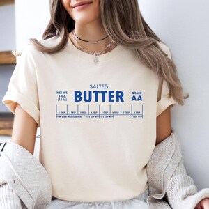 May include: A white t-shirt with a blue graphic that reads "SALTED BUTTER GRADE AA" and includes a measurement guide for tablespoons and cups. The graphic is a stylized butter stick with measurements for 1, 2, 3, 4, 5, 6, and 7 tablespoons. The graphic also includes measurements for 1/4 cup, 1/3 cup, and 1/2 cup. The t-shirt is a light cream colour and has a crew neck.