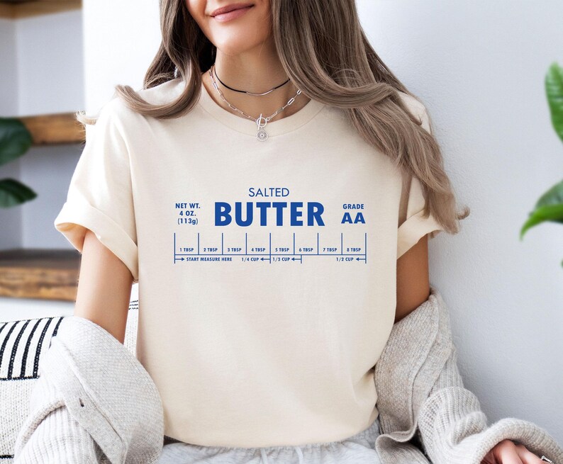 May include: A white t-shirt with a blue graphic that reads "SALTED BUTTER GRADE AA" and includes a measurement guide for tablespoons and cups. The graphic is a stylized butter stick with measurements for 1, 2, 3, 4, 5, 6, and 7 tablespoons. The graphic also includes measurements for 1/4 cup, 1/3 cup, and 1/2 cup. The t-shirt is a light cream colour and has a crew neck.