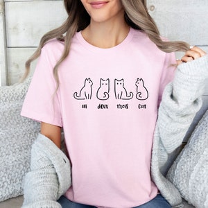 Peut inclure: Un t-shirt rose clair avec un dessin au trait noir de quatre chats. Le texte "un deux trois Cat" est en dessous des chats. Le t-shirt est fait d'une mati&egrave;re douce.