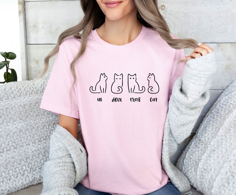 Peut inclure: Un t-shirt rose clair avec un dessin au trait noir de quatre chats. Le texte "un deux trois Cat" est en dessous des chats. Le t-shirt est fait d'une mati&egrave;re douce.