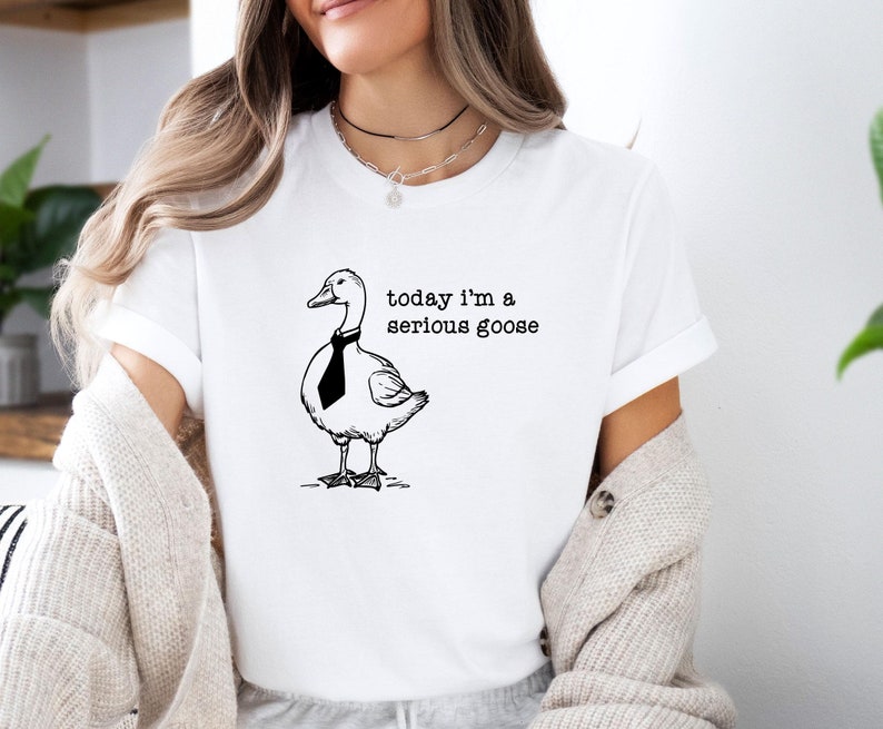 Pu&ograve; includere: Maglietta bianca con un'illustrazione in bianco e nero di un'oca con una cravatta. Il testo "today i'm a serious goose" &egrave; stampato sotto l'oca.