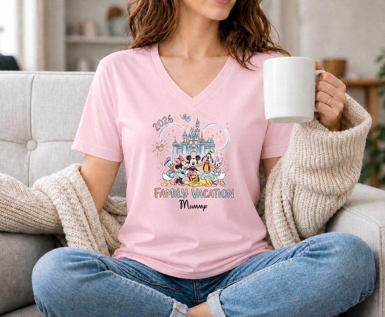 May include: A pale pink V-neck t-shirt with a Disney-themed graphic. The design includes a castle, Mickey Mouse, Minnie Mouse, Donald Duck, and Goofy, with the text "2026 Family Vacation Mummy".