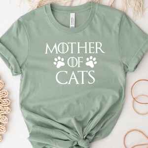 May include: A light green t-shirt with a white graphic that says "Mother of Cats" with paw prints on either side of the word "of".