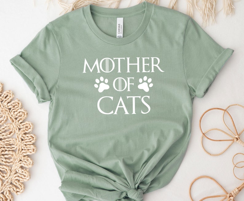 May include: A light green t-shirt with a white graphic that says "Mother of Cats" with paw prints on either side of the word "of".