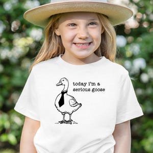Pu&ograve; includere: Una maglietta bianca con un'illustrazione in bianco e nero di un'oca con una cravatta. Il testo "today i'm a serious goose" &egrave; stampato sulla maglietta.
