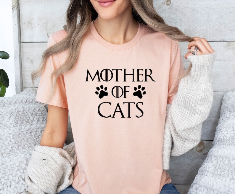 May include: A pink t-shirt with the text "Mother of Cats" in black, featuring a paw print design.