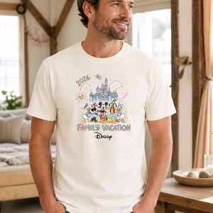 May include: Cream-colored t-shirt featuring a colorful graphic of Disney characters in front of a castle. The text "2026" and "Family Vacation Daddy" are also displayed. The shirt is short-sleeved.