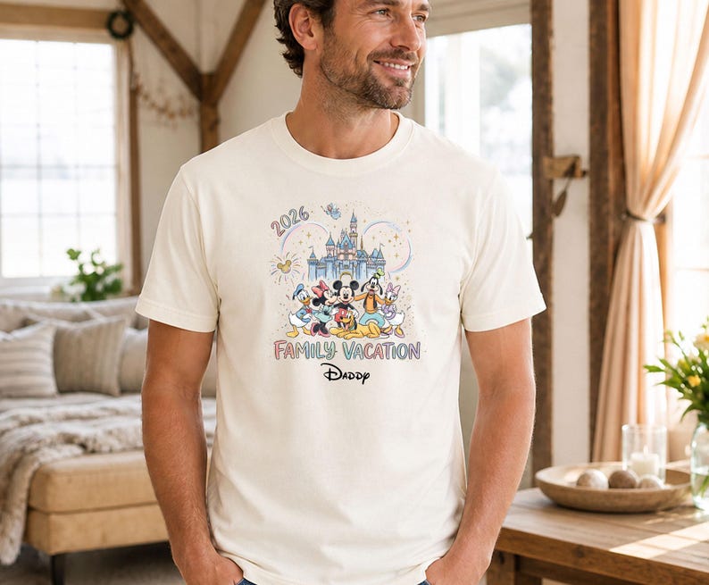 May include: Cream-colored t-shirt featuring a colorful graphic of Disney characters in front of a castle. The text "2026" and "Family Vacation Daddy" are also displayed. The shirt is short-sleeved.