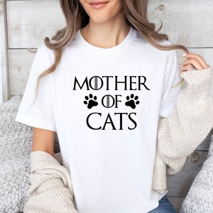 May include: White t-shirt with black text that reads "Mother of Cats" with two paw prints on either side of the word "of".