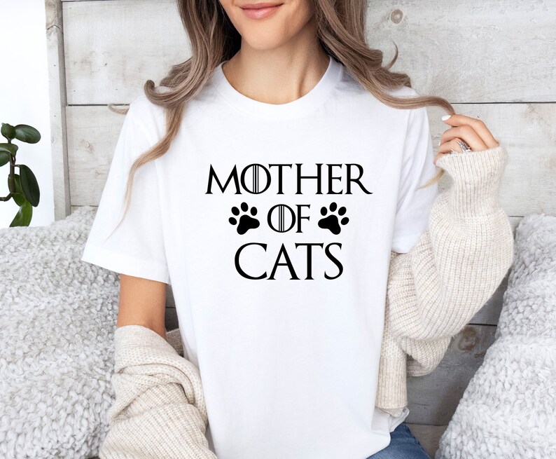 May include: White t-shirt with black text that reads "Mother of Cats" with two paw prints on either side of the word "of".