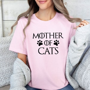 May include: A pink t-shirt with the text "Mother of Cats" in black lettering. The text is styled to look like the Game of Thrones title card, with a paw print replacing the crown.