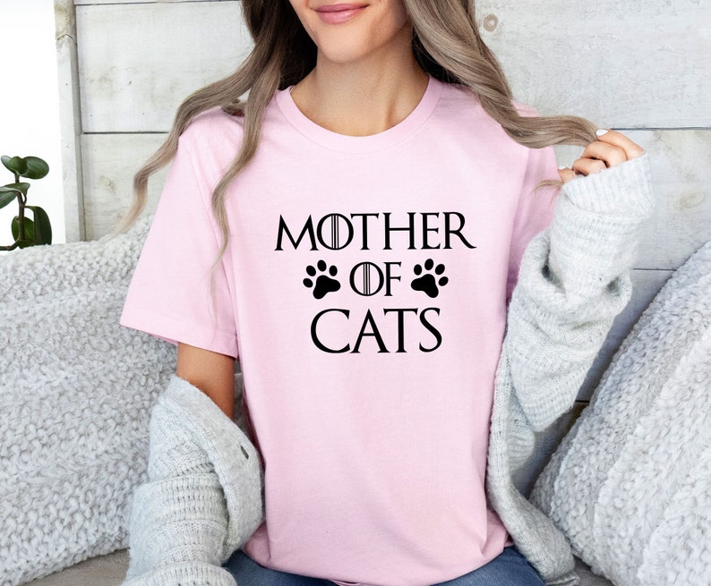 May include: A pink t-shirt with the text "Mother of Cats" in black lettering. The text is styled to look like the Game of Thrones title card, with a paw print replacing the crown.
