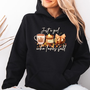May include: A black hooded sweatshirt featuring a graphic print of a coffee mug with a leaf design, a candle labelled cozy autumn, and a pumpkin with a bow, surrounded by fall leaves, with the text Just a girl who loves fall printed in white.