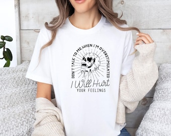 May include: A white short-sleeved t-shirt featuring a black graphic of a melting skull surrounded by radiating lines. The text reads: DON'T TALK TO ME WHEN I'M OVERSTIMULATED I Will Hurt YOUR FEELINGS. The design is centered on the chest area.