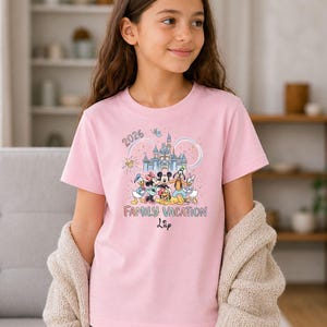 May include: A light pink t-shirt featuring a colorful graphic of Disney characters in front of a castle. The shirt has the text "2026 Family Vacation" and a name. The shirt is short-sleeved.