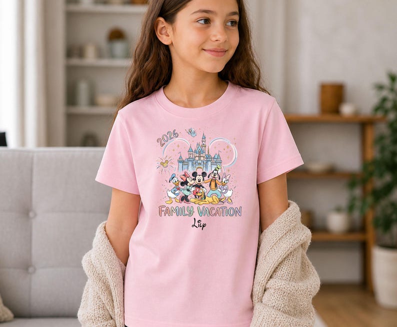May include: A light pink t-shirt featuring a colorful graphic of Disney characters in front of a castle. The shirt has the text "2026 Family Vacation" and a name. The shirt is short-sleeved.