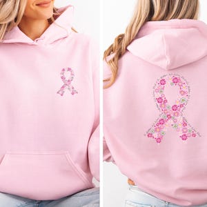 Könnte beinhalten: Ein hellrosa Kapuzenpullover mit einer kleinen floralen Awareness-Schleife auf der linken Brust und einer großen floralen Schleife auf der Rückseite, die den Text In October We Wear Pink entlang der Schleifenform eingearbeitet hat.
