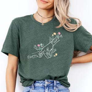 May include: Dark green t-shirt featuring a white line drawing of two hands holding each other, with the word "Mummy" and "Name" in cursive. The design includes floral accents in pink, yellow, and green.