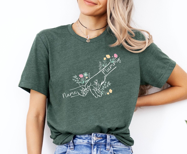 May include: Dark green t-shirt featuring a white line drawing of two hands holding each other, with the word "Mummy" and "Name" in cursive. The design includes floral accents in pink, yellow, and green.