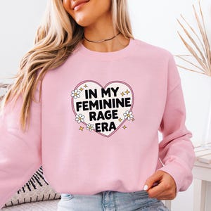 Könnte beinhalten: Ein hellrosa Sweatshirt mit Rundhalsausschnitt und einem zentralen Herz-Grafikmotiv auf der Brust, das den schwarzen Text "IN MY FEMININE RAGE ERA" enthält, umgeben von kleinen weißen Gänseblümchen und gelben vierzackigen Sternsymbolen.