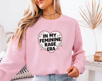Könnte beinhalten: Ein hellrosa Sweatshirt mit Rundhalsausschnitt und einem zentralen Herz-Grafikmotiv auf der Brust, das den schwarzen Text "IN MY FEMININE RAGE ERA" enthält, umgeben von kleinen weißen Gänseblümchen und gelben vierzackigen Sternsymbolen.