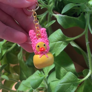 May include: A yellow rubber ducky with a pink bunny costume. The ducky has a red bow and a gold chain attached to its head.