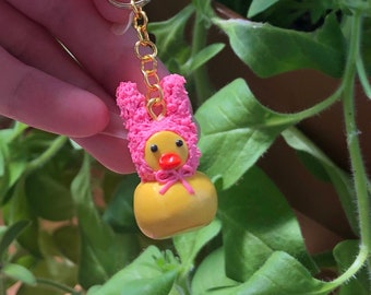 May include: A yellow rubber ducky with a pink bunny costume. The ducky has a red bow and a gold chain attached to its head.