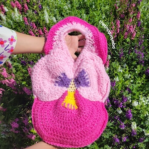 May include: A crocheted pink, purple and yellow pansy flower-shaped bag with a handle. The bag is held in front of a background of purple and white flowers.