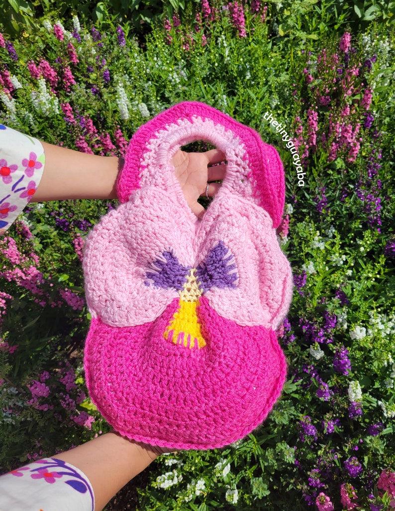 May include: A crocheted pink, purple and yellow pansy flower-shaped bag with a handle. The bag is held in front of a background of purple and white flowers.