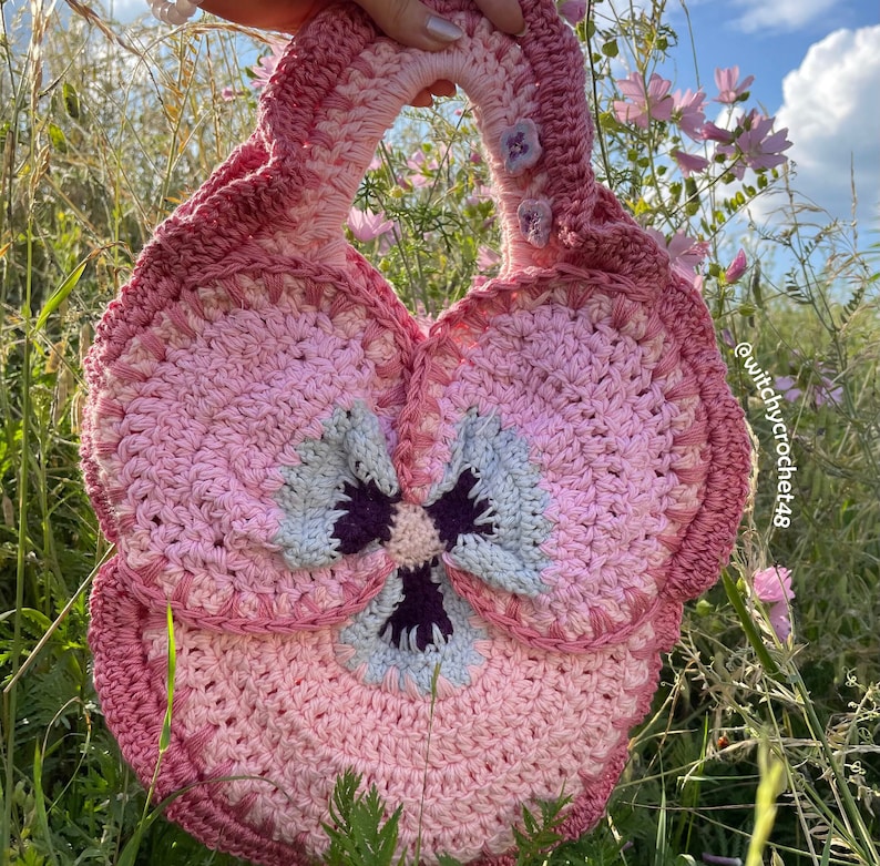 May include: A pink crocheted handbag shaped like a pansy flower with a purple and blue centre. The bag has a handle and is decorated with small pink buttons.