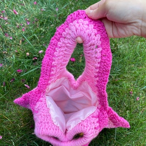 May include: A pink crocheted bag with a heart-shaped opening and a pink satin lining. The bag is made with a gradient of pink yarn, from a lighter pink to a darker pink.