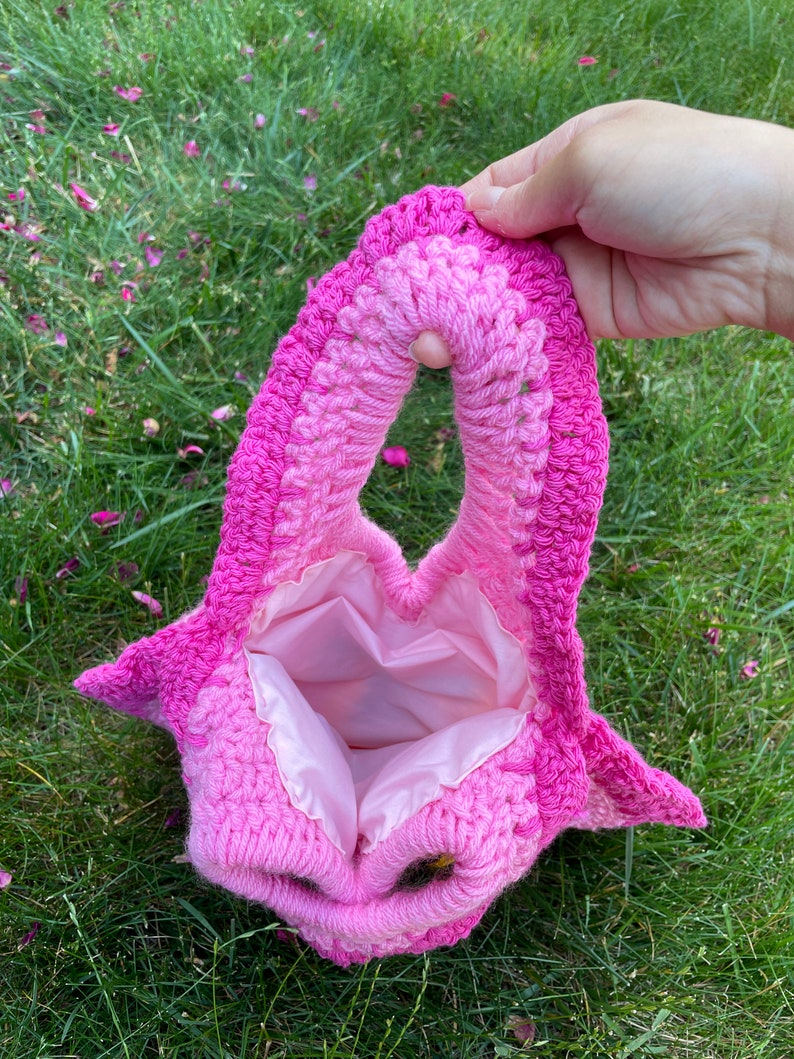 May include: A pink crocheted bag with a heart-shaped opening and a pink satin lining. The bag is made with a gradient of pink yarn, from a lighter pink to a darker pink.