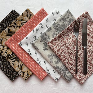 Peut inclure: Ensemble de serviettes en tissu pliées avec divers motifs, dont des motifs floraux, géométriques et toiles de Jouy. La palette de couleurs comprend le noir, l'or, le rouge et le blanc. Une fourchette et un couteau reposent sur une serviette fleurie.