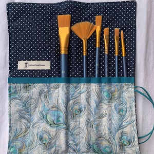 Könnte beinhalten: Eine teal- und weiße Stoffrolle mit einem Pfauenfeder-Design. Die Rolle hat eine schwarze, weiß gepunktete Stoffklappe mit einem Etikett, auf dem "Old Town Creations" steht. Die Rolle enthält sechs Pinsel mit goldenen Griffen.