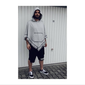 May include: A gray hooded poncho with a fringe trim and a pocket. The person is wearing black shorts and black and white sneakers.