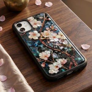 May include: Black smartphone case with a floral design featuring white blossoms and dark branches. The case is on a wooden table, with a small vase of pink flowers and scattered petals nearby.