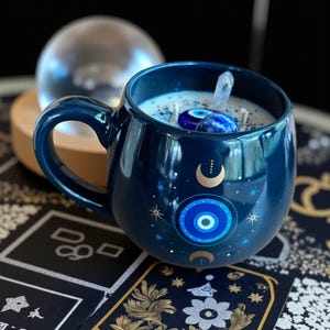 Op de afbeelding: Een donkerblauwe keramische mokkaars met een witte was binnenkant, versierd met hemelse en boze oogontwerpen. De kaars is versierd met kristallen en blauwe edelstenen. Een heldere glazen bol staat op de achtergrond.