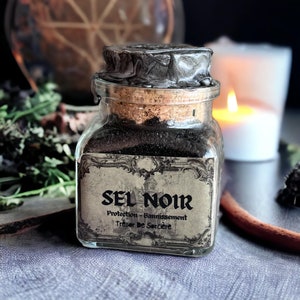 Peut inclure: Un bocal en verre avec un couvercle en liège noir et une étiquette qui indique "Sel Noir Protection - Bannissement Trésor De Sorcière". Le bocal est rempli de sel noir.