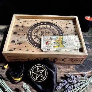 Può includere: Una scatola rettangolare in legno con un design di sole e luna inciso al laser sul coperchio, contenente un mazzo di tarocchi. Un sacchetto nero con coulisse con un simbolo di pentagramma dorato e un mazzetto di lavanda secca sono davanti.