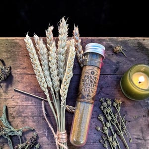 Puede incluir: Un vial de vidrio con tapa plateada y una etiqueta con texto en francés: "Poudre des Chemins Ouverts, Poudre pour la Chance" se encuentra sobre una superficie de madera, junto a un manojo de trigo y una vela verde encendida.